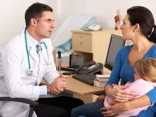 A mother with her daughter sitting on her knee while she talks to a concerned-looking doctor.