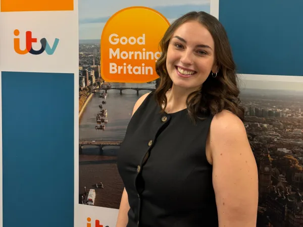 A young woman in her early 20s with brown hair, smiling and wearing a short-sleeve black shirt, stands in front of an 'ITV Good Morning Britain' backdrop.