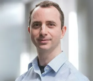 A man is smiling in a headshot, he has short hair and wearing a light blue shirt. 