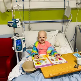 A young girl with a bald head sits in a hospital bed, smiling. She wears a colourful rainbow-patterned dressing gown, with a bright colouring book open in front of her.