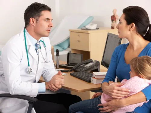 A mother with her daughter sitting on her knee while she talks to a concerned-looking doctor.