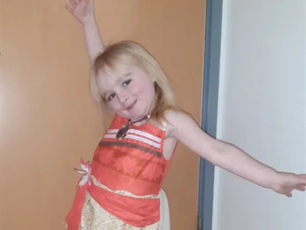 A smiling young girl with blonde hair stands with her arms outstretched, wearing an orange top and a beige skirt. 