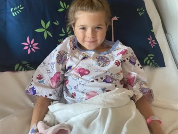 A young girl with brown hair styled in a braid lies in a hospital bed, smiling.