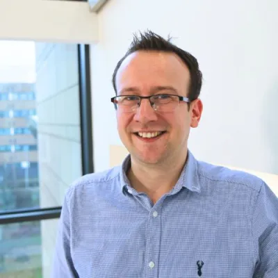 Tim wearing glasses and smiling at the camera. He has short brown hair and is wearing a pale blue shirt.