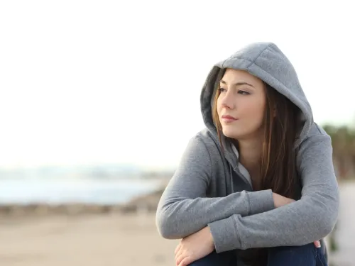 A young woman in a grey hoodie, sitting on a beach and looking thoughtful.